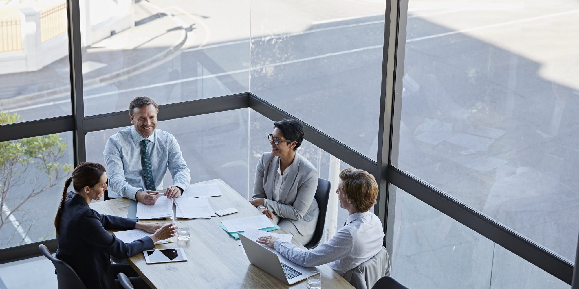 Business people at conference table in modern office High angle view of smiling business people in meeting. Male and female professionals are sitting at conference table against window. They are working in office.