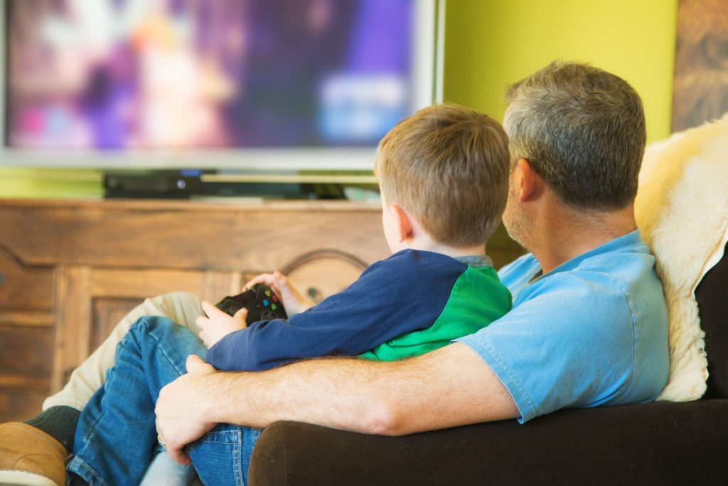 Rear view of mature father teaching a video game to his young son sitting on his lap